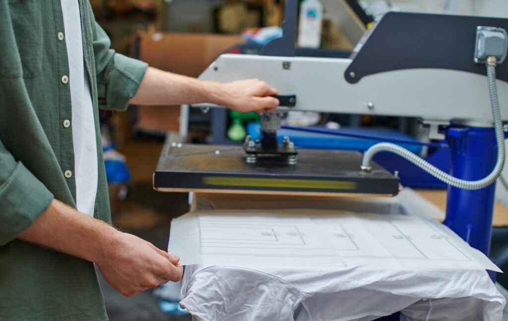 Cropped view of craftsman holding layer on t-shirt and working with screen printing machine